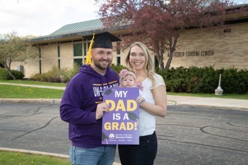 A male student in a grad cap posed with his wife and child outside to celebrate his masters degree..