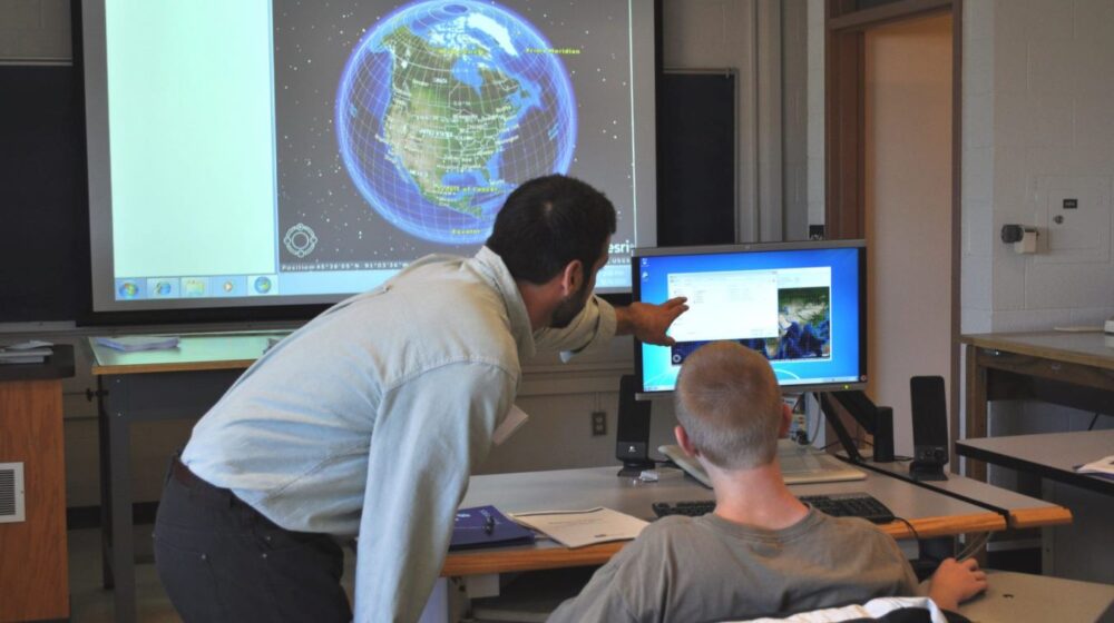 Faculty member teaching student at computer screen with the world image on the screen projector.